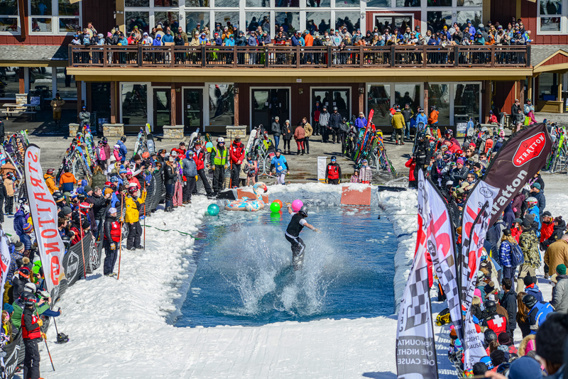 A person riding a snowboard, skims across a pool of water at the bottom of a snow-covered ski slope.