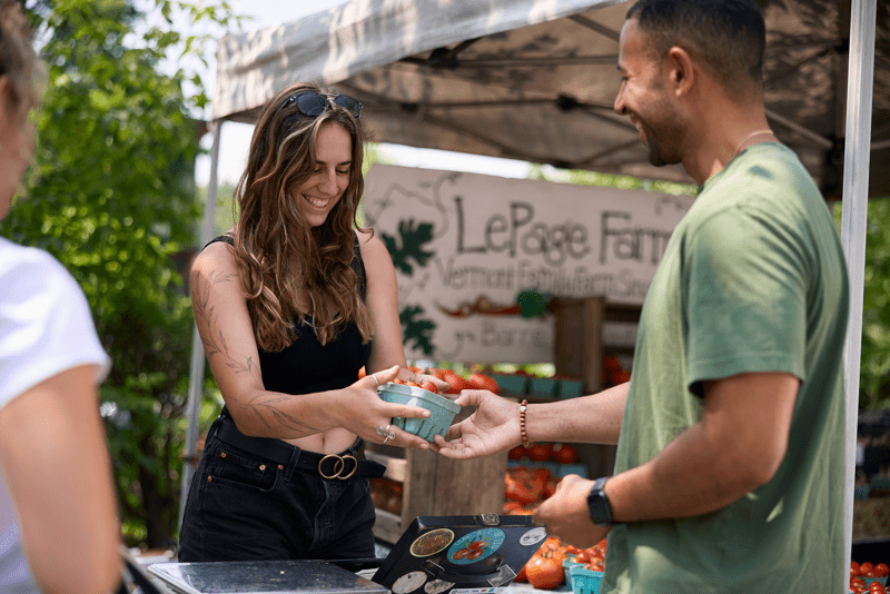Two people at an outdoor farmers market smiling. One is handing the other a container of tomatoes. A third person is partially visible on the left. 