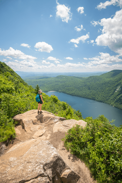 A person stands on an exposed rock ledge to look out over a mountain valley and lake below on a warm sunny day.