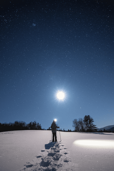 A person snowshoes by moonlight wearing a headlamp.