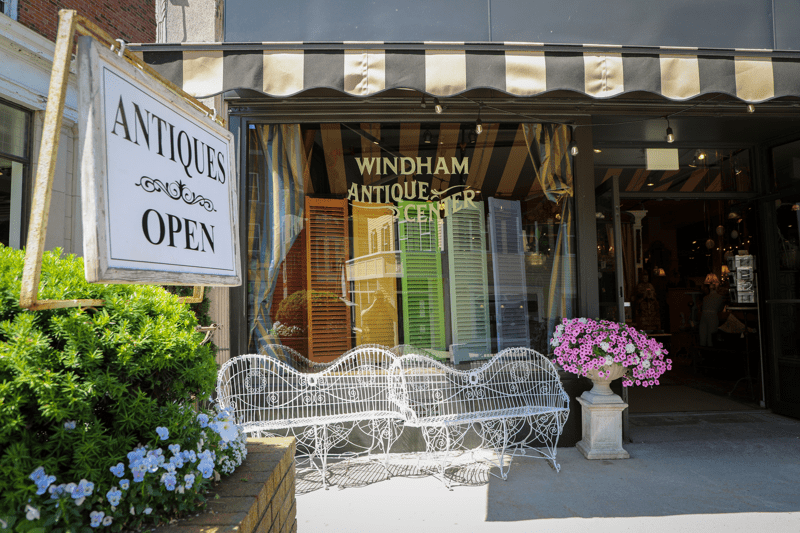 A sign reading “Antique Shop” and “Open” in front of a white building seen from the outside in the summer. 