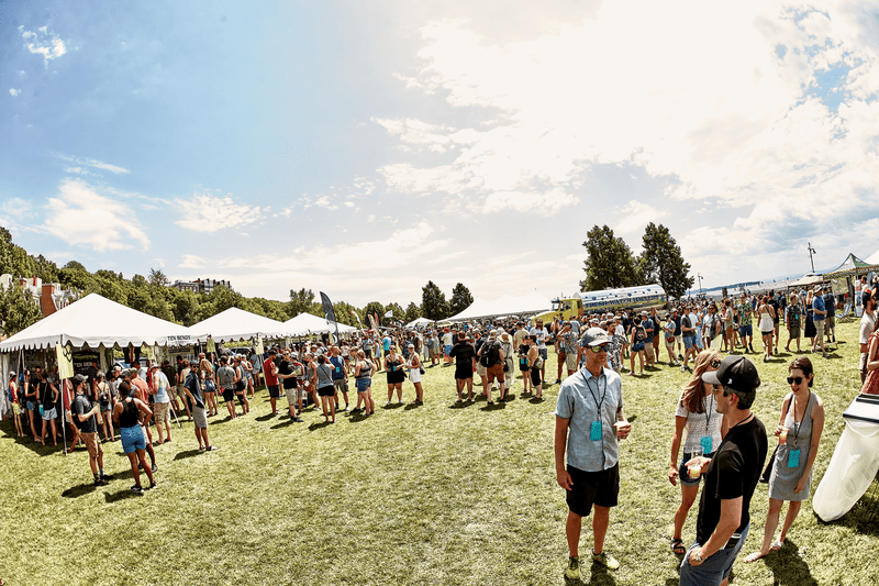 A crowd of people stands around at an outdoor festival on a sunny day.