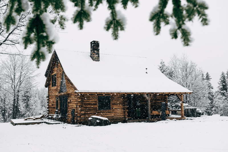 A log cabin seen from outdoors in the winter as snow falls. It’s surrounded by woods.