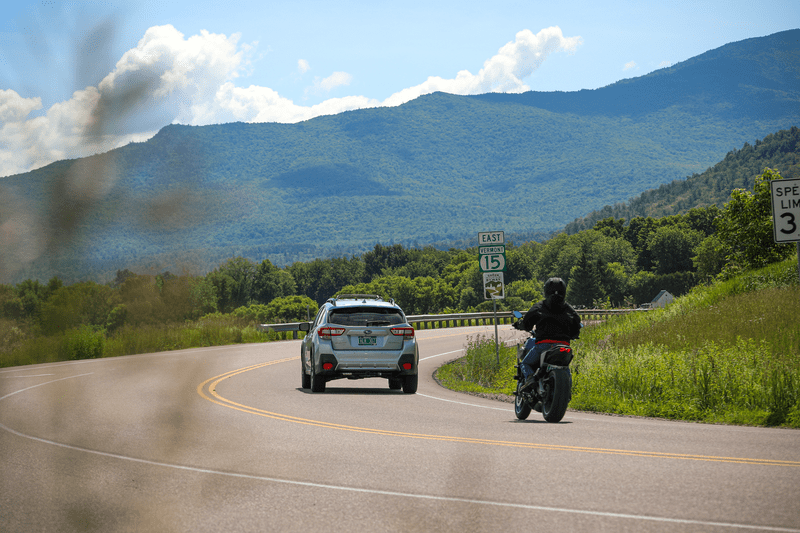 A car and a motorcycle travel along a road with a mountain behind.
