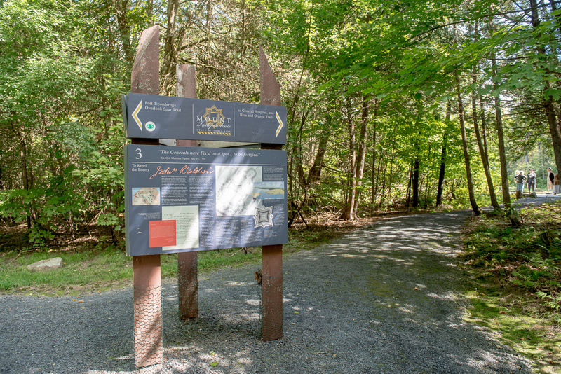 A sign outlining offerings at Mount Independence State Historic Site next to a trail in the woods on a sunny day.