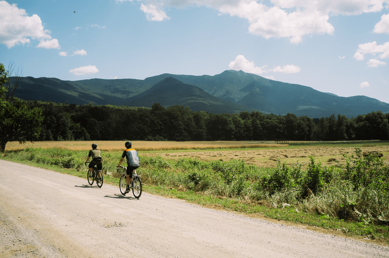 Two people ride bicycles along a gravel road on a sunny day.