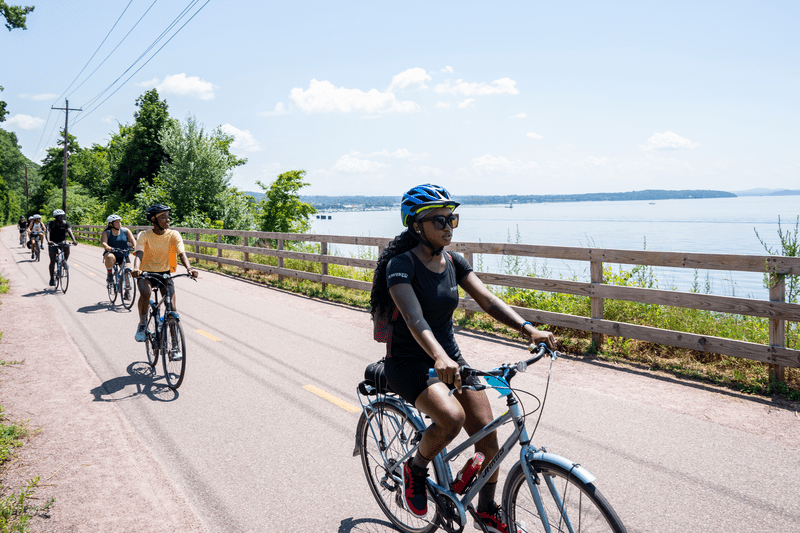 A group of people ride bicycles on a path along a large body of water on a sunny day.