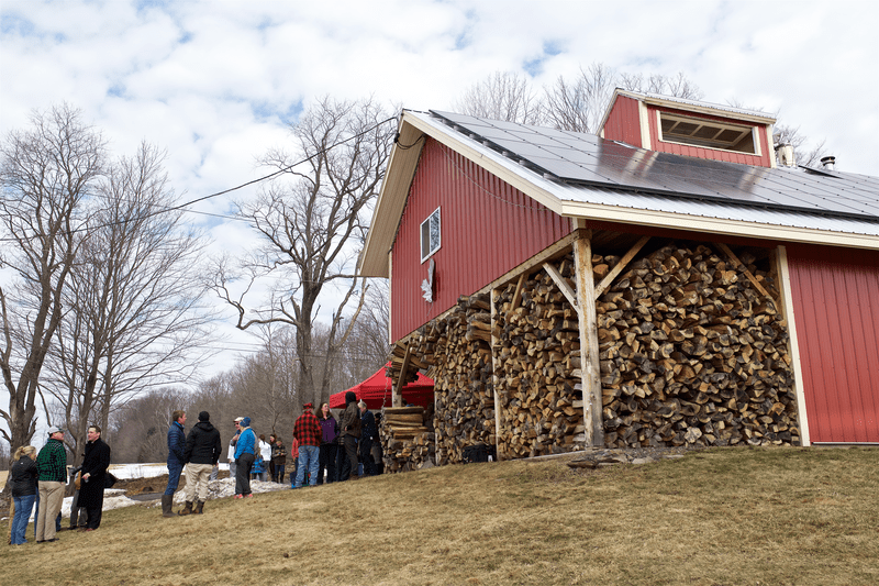 A small group of people stand by the entrance to a red barn filled with split wood.