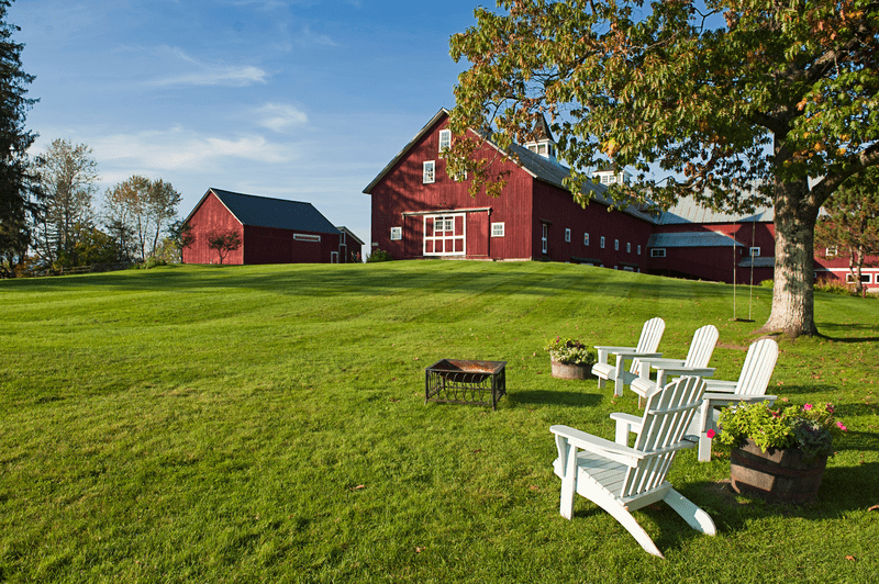 White Adirondack chairs surround a fire ring on a green lawn with a red barn in the background.