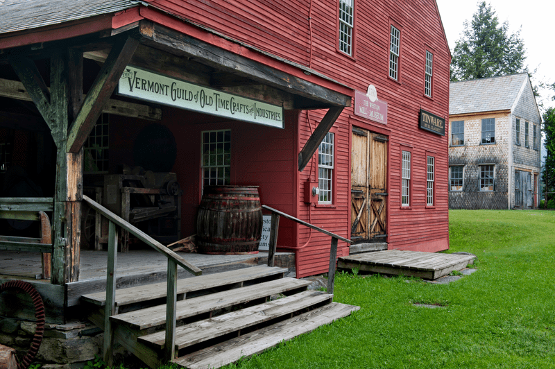 A red building has stairs leading to a door and a sign above that reads Vermont Guild of Old Time Crafts and Industries.