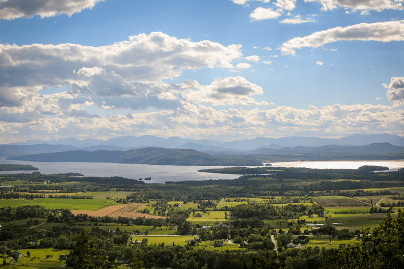 The view from the top of a Vermont mountain, with green fields and buildings visible below and other mountains in the background.