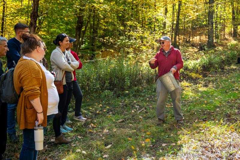 A guide holding a metal bucket speaks to a small tour group along a grassy trail in a sunny autumn forest.