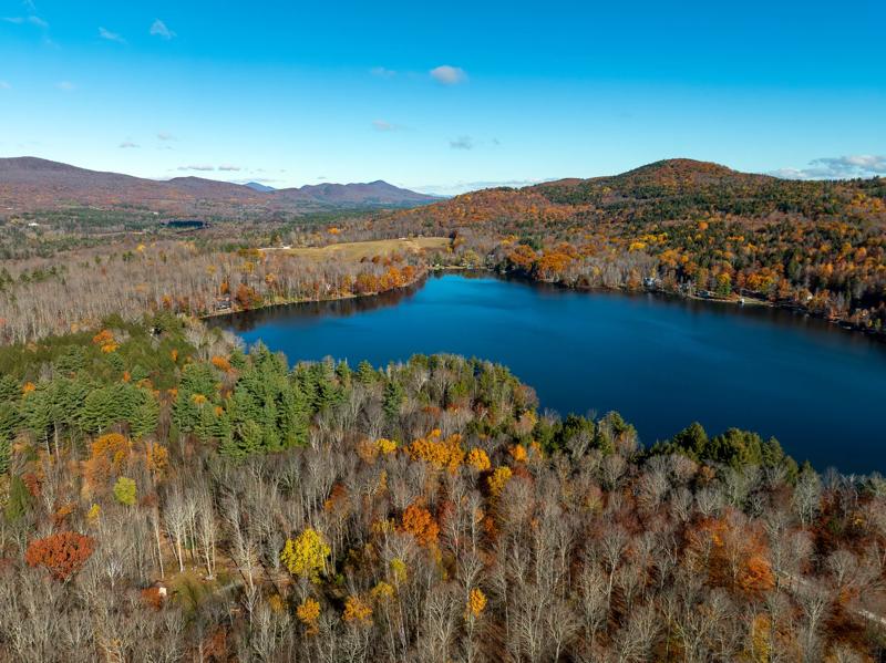 An aerial view of a lake in the late fall. Most of the trees have lost their leaves, though a few still hold onto red, orange, or yellow color.