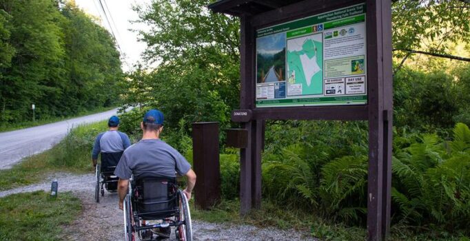 Two individuals visit a wheelchair accessible trail with a large trailhead sign by a roadside.