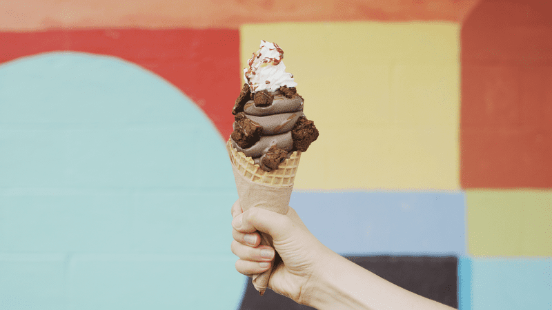 A hand holds a chocolate soft ice cream in a waffle cone with toppings in front of a colorful wall.
