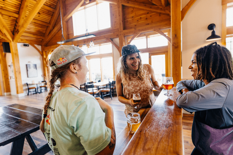 Three people stand and talk inside a brewery. Beer glasses sit on a wood counter in between the people.