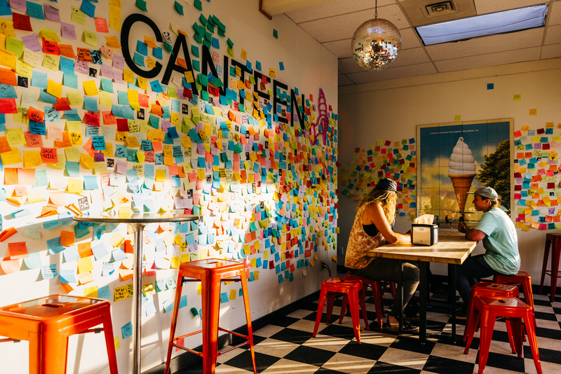 Two people sit at a table in an ice cream shop with a disco ball on the ceiling. Colorful sticky notes with handwritten messages line one wall.
