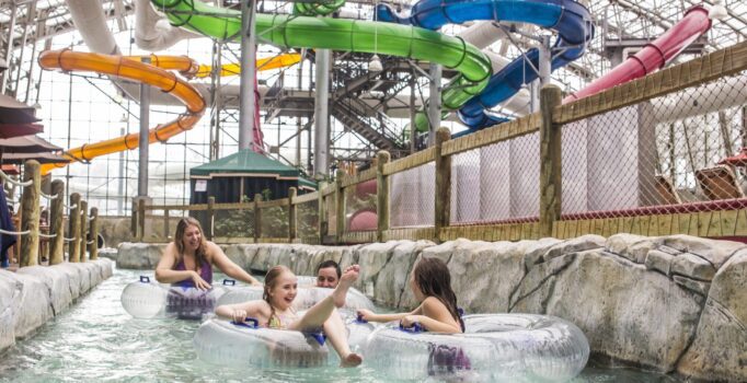 Four people swim in innertubes with colorful waterslides behind them at an indoor water park.