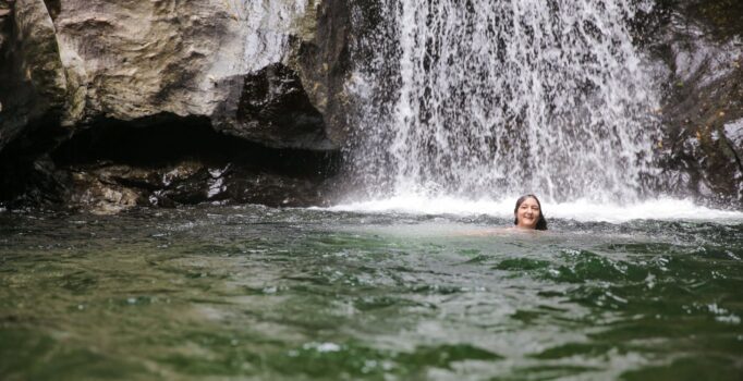 A person swims in front of a waterfall.