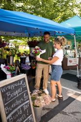 People standing under a vendor tent at a farmers market looking at potted flowers.