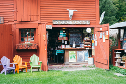 A red barn has an open door exposing items inside and Adirondack chairs outside on a sunny day.