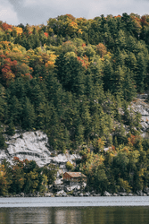 Seen from afar, a building is built on the shore of a lake with exposed rocks and surrounded by trees with fall colors.