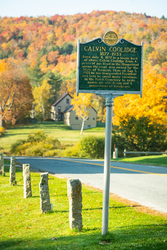 A green historic marker sign standing on a trail with autumn foliage behind.