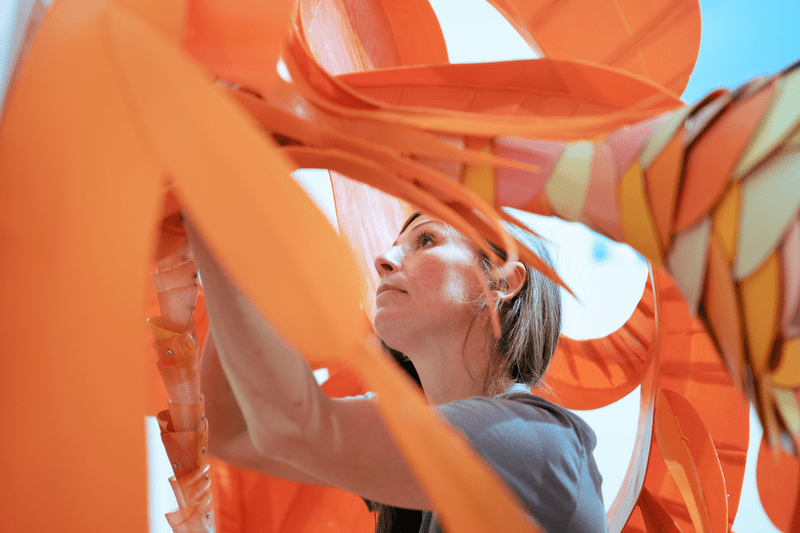 Seen from below, a person stands within a curved, orange sculpture of orange