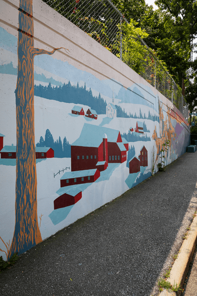 A wall next to a sidewalk that's painted with a snowy scene and red buildings and trees.