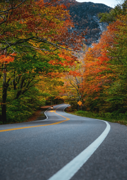 A paved road winds through a mountain valley filled with autumn-colored trees.