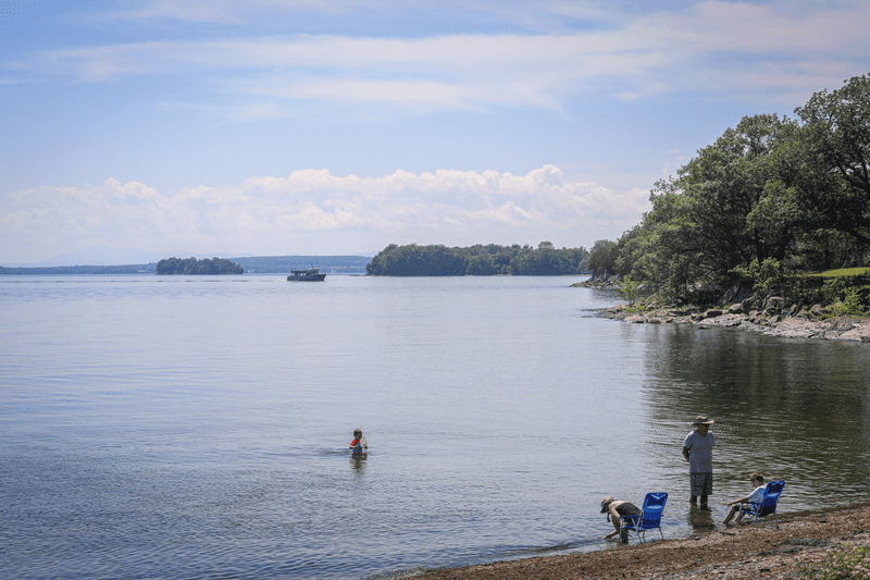 A person swims while three others sit on the beach at a quiet lake.