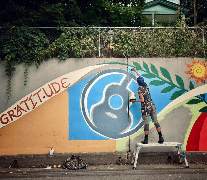 Seen from behind, a person paints a mural that includes an acoustic guitar, lots of colors, and the word “gratitude.”