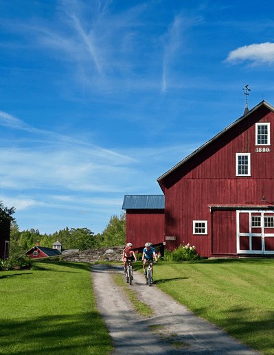 Two people ride their bikes along a dirt path in front of a red barn.