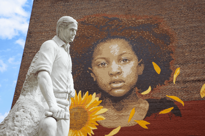 A brick wall with a painting of a person and flowers and a white marble sculpture in a downtown in the summer.