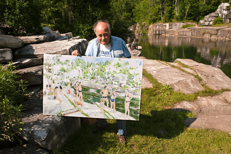 A person stands in grass holding up a painted piece of art. A pond can be seen in the background.