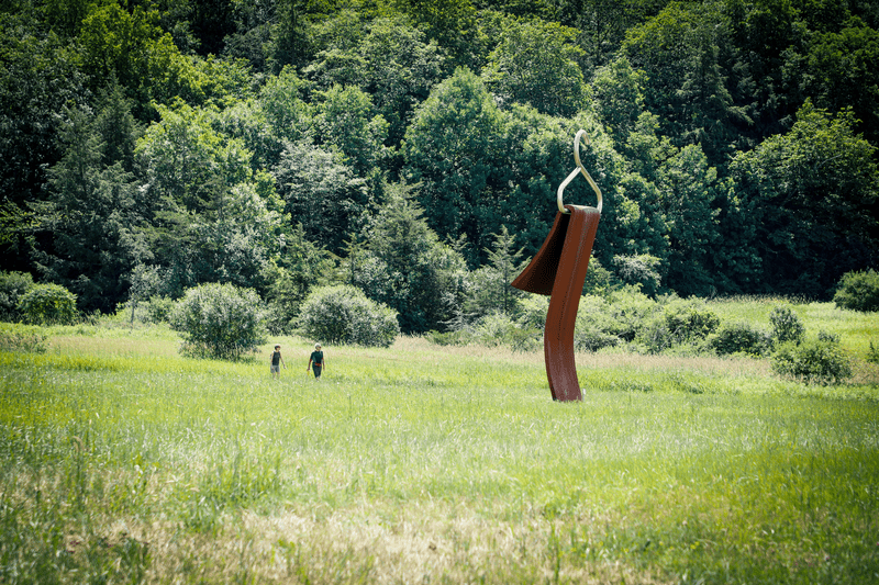 Two people walk in grass near a tall sculpture with a clothes hanger on top.