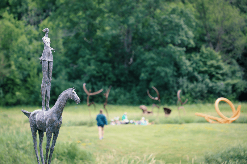 A metal sculpture of a person standing on a horse. Other sculptures and people walking can be seen in the background.