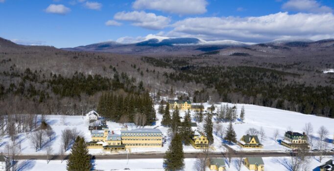 Seen from above, a wintery mountain valley and a series of yellow buildings along a country road.