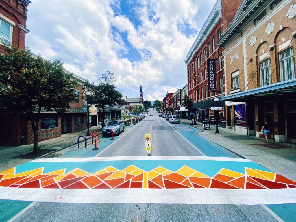 A colorful, geometric crosswalk on the street in front of a row of historic brick buildings.