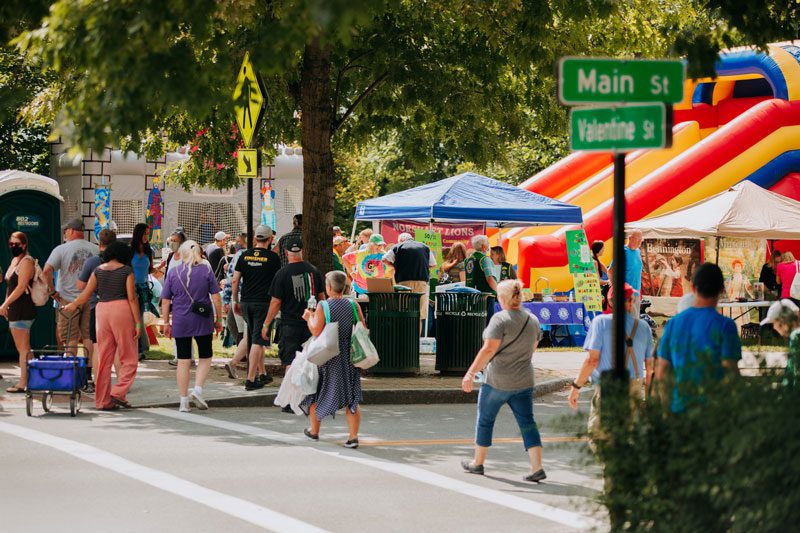 People crowd around a festival, including a colorful bounce house and vendor stalls, in a bustling downtown.  