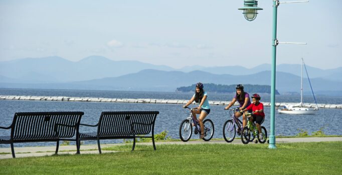 Three people ride bikes along a waterfront path with mountains in the background on a sunny, warm day.