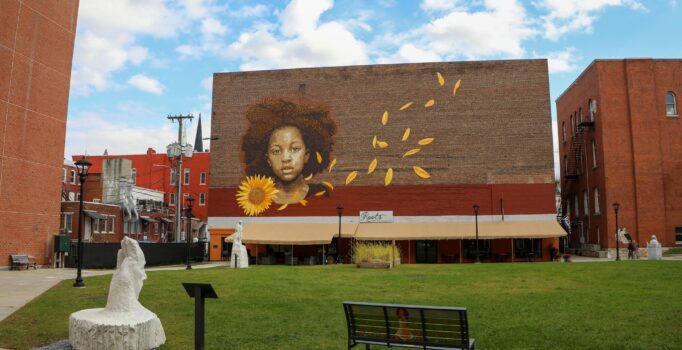 A town green surrounded by marble sculptures and brick buildings. A mural of a child with a sunflower is painted on a brick building.