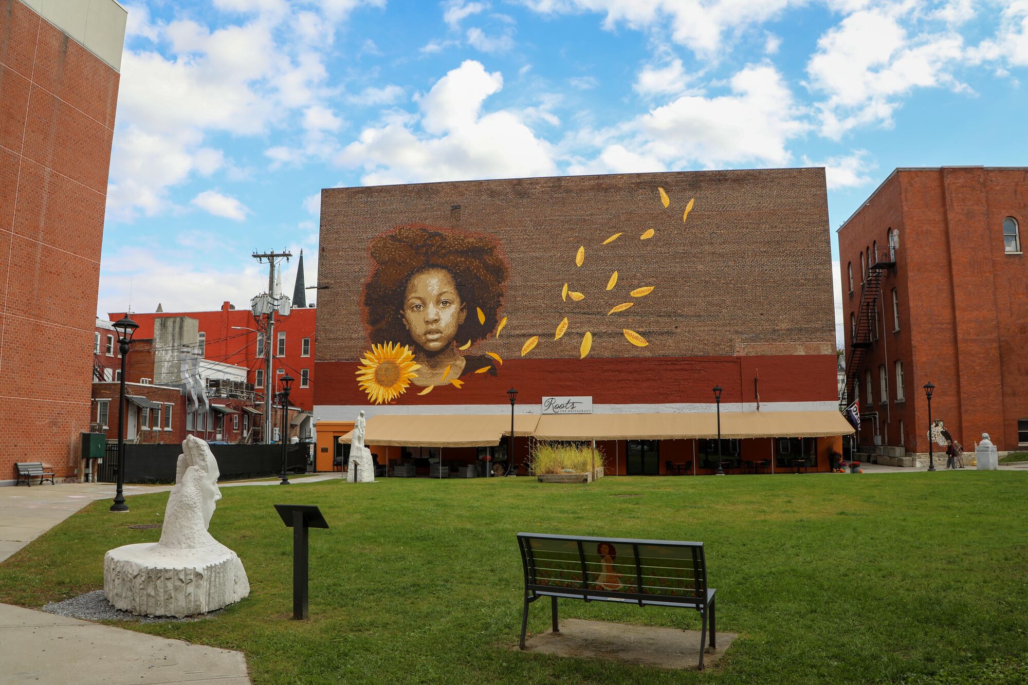 A town green surrounded by marble sculptures and brick buildings. A mural of a child with a sunflower is painted on a brick building.