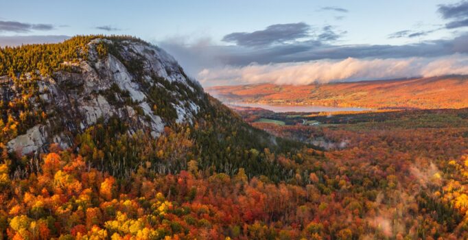 Seen from above, a lake surrounded by mountain peaks with bright red, orange, and yellow leaves on the trees in the fall.
