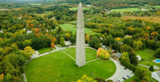 Seen from above, a tall stone monument surrounded by grass, trees, and mountains.