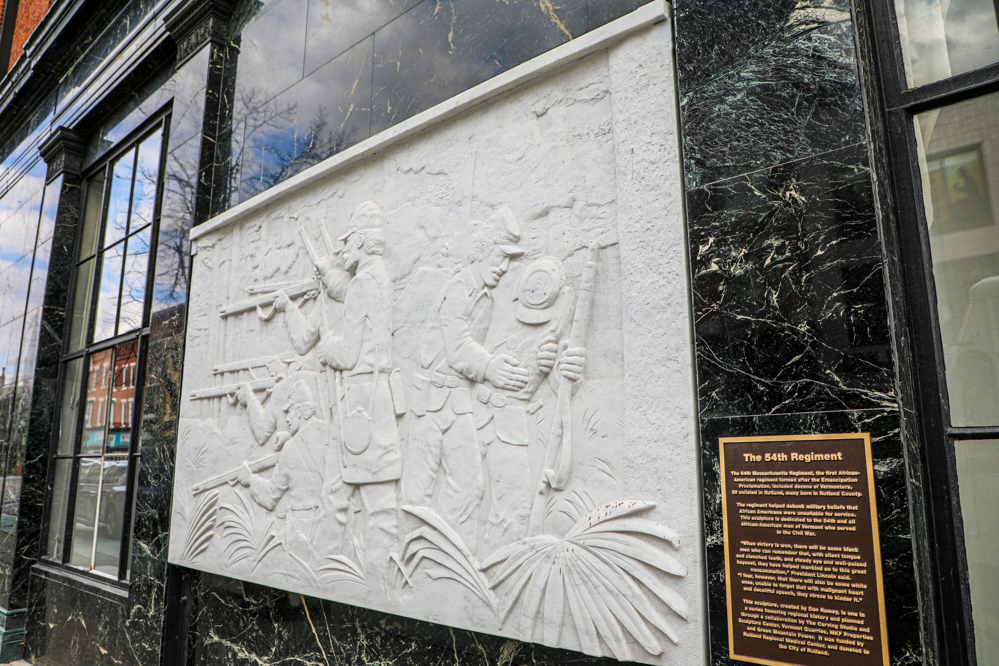 A white marble bas-relief sculpture depicting Union soldiers holding rifles mounted on a dark marble building facade, accompanied by an informational plaque titled "The 54th Regiment."
