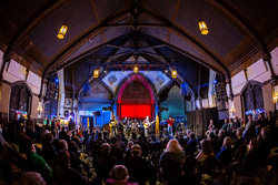 View of a large, indoor hall with a stage in front and a crowd watching a performance.