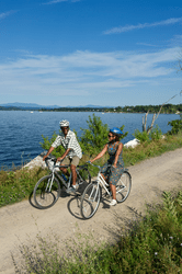 Two people ride bicycles along a gravel path close to water.