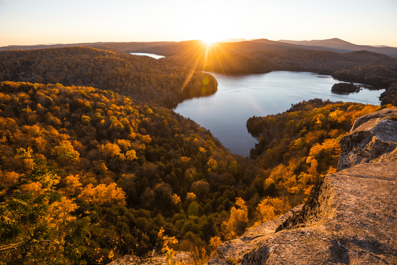 A lake seen from a mountain ledge with bright red, yellow, and orange leaves on the trees seen from above and a distance. The sun peeks out in the distance.