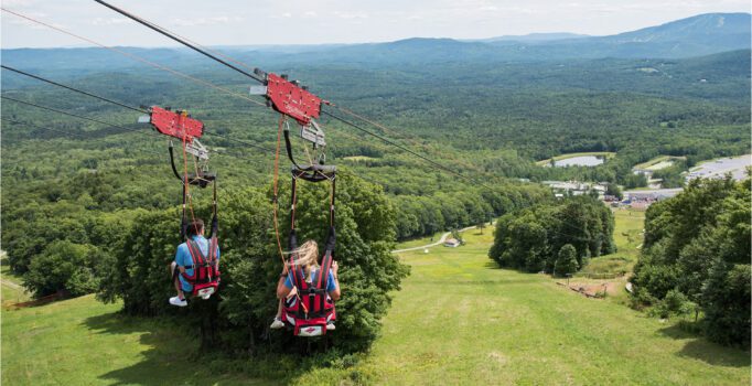 Two people are riding a zipline over a ski slope in the summer.